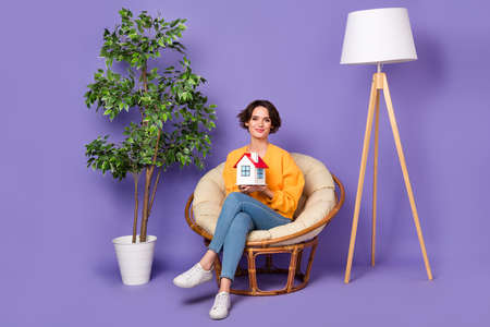 Portrait of attractive cheerful girl sitting in chair holding in hands paper home isolated over violet lilac color backgroundの写真素材