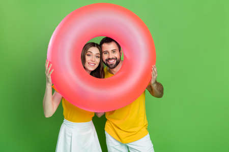 Portrait of two cheerful positive people hands hold look through inflatable ring isolated on green...