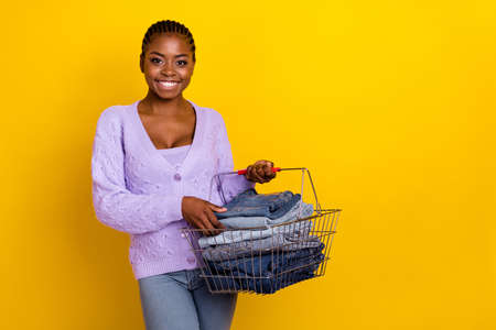 Photo of cheerful pretty girl hands hold shopping basket stack denim jeans isolated on yellow color backgroundの写真素材