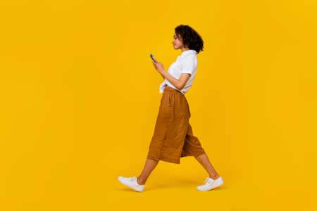 Full length profile portrait of pretty nice lady walk hold use telephone chatting isolated on yellow color backgroundの写真素材