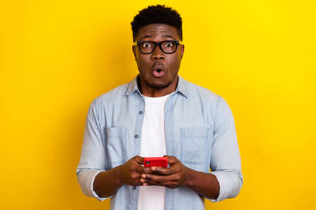 Photo of impressed young brunet guy hold telehone wear eyewear jeans shirt isolated on yellow color...