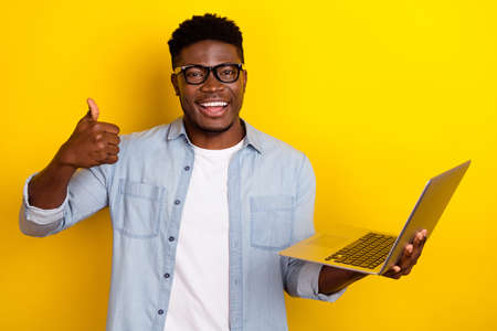 Photo of funky young brunet guy hold laptop thumb up wear eyewear jeans shirt isolated on yellow...