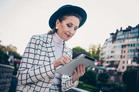 Photo of pretty adorable woman wear plaid jacket cap typing modern gadget enjoying sunshine outside city residential complexの写真素材