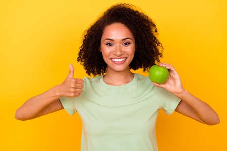 Photo of confident cute lady wear green t-shirt rising apple white teeth thumb up isolated yellow color backgroundの写真素材