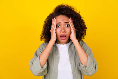 Photo of stressed curly young brunette lady hold head wear grey shirt isolated on vivid yellow color backgroundの写真素材