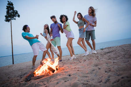 Full length photo of millennial guys ladies dance near fire wear casual cloth by the ocean