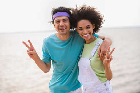 Photo of pretty charming young husband wife embracing smiling showing v-signs outside seaside beach