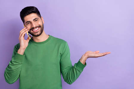Photo of cheerful good mood handsome guy dressed green pullover showing empty space talking on phone isolated on violet color backgroundの写真素材