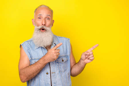 Photo of funny impressed man pensioner dressed denim vest pointing fingers empty space isolated yellow color backgroundの写真素材