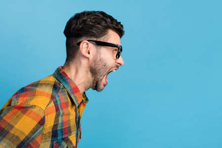 Side profile photo of angry stylish millennial guy in spectacles dressed checkered shirt loud screaming isolated on blue color backgroundの写真素材