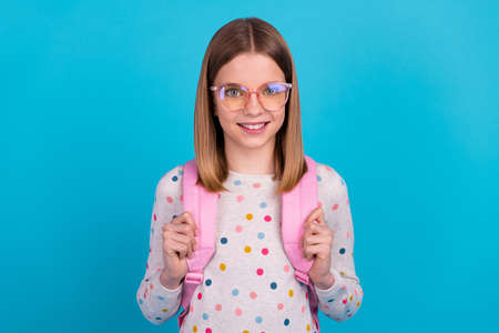 Photo of confident adorable small girl wear dotted shirt eyeglasses backpack smiling isolated blue color backgroundの写真素材