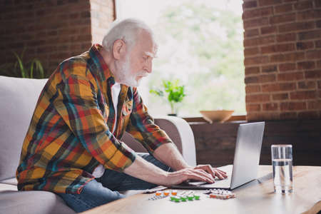 Photo of lonely busy man pensioner wear checkered shirt typing modern gadget sitting sofa indoors house roomの写真素材