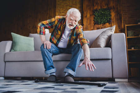 Photo of depressed upset retired man dressed plaid shirt falling nursery cane trying getting up couch indoors apartmentの写真素材