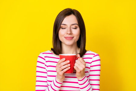 Photo portrait of adorable dreamy young woman smell coffee flavor enjoy break wear stylish striped look isolated on yellow color backgroundの写真素材