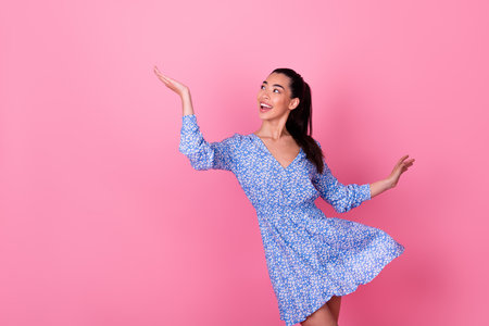 Portrait of young happy stylish lady wear blue dress showing new advertisement look empty space isolated on pink color backgroundの写真素材