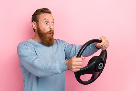 Portrait of staring astonished guy with ginger beard blue pullover hold steering wheel look empty space isolated on pink color backgroundの写真素材