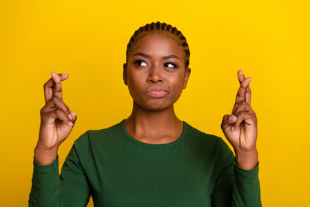 Photo of doubtful unsure young lady dressed green shirt fingers crossed looking empty space isolated yellow color backgroundの写真素材