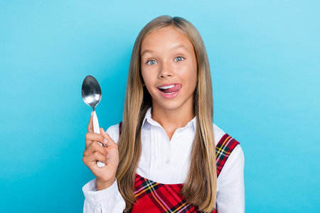 Closeup photo of young little cute schoolgirl hold spoon tongue out hungry want eat soup isolated on blue color backgroundの写真素材