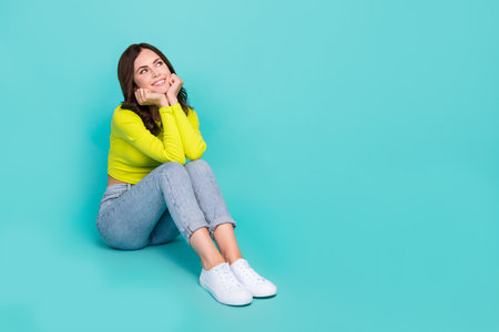 Full body portrait of cheerful pretty lady sit floor hands touch cheekbones look empty space isolated on teal color backgroundの写真素材