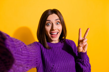 Portrait of charming astonished girl make selfie demonstrate v-sign isolated on yellow color backgroundの写真素材