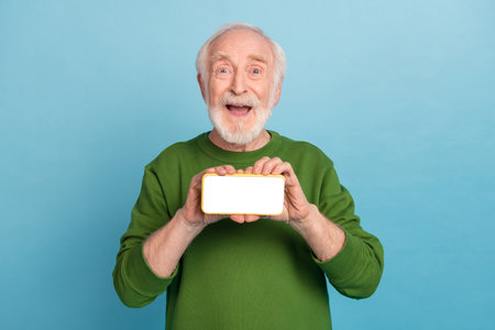 Portrait of astonished cheerful aged man hold telephone demonstrate empty space screen isolated on blue color backgroundの写真素材