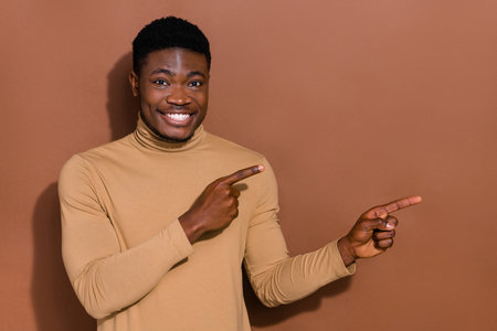 Photo of funny millennial brunet guy indicate empty space wear beige turtleneck isolated on brown color backgroundの写真素材