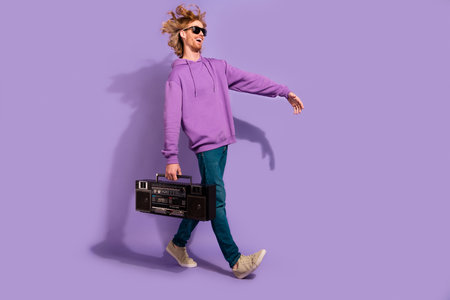 Full size photo of funky beard young guy go with boombox wear sunglass hoodie jeans boots isolated on violet backgroundの写真素材