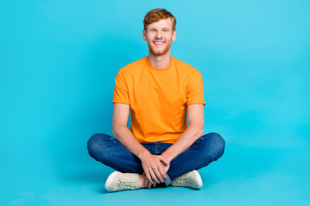 Full length photo of funky attractive man wear orange t-shirt sitting floor legs crossed isolated teal color backgroundの写真素材