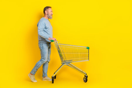 Full length profile portrait of aged person push shopping car look empty space isolated on yellow color backgroundの写真素材