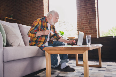 Photo of depressed sad retired man dressed plaid shirt sitting couch reading pills prescription...