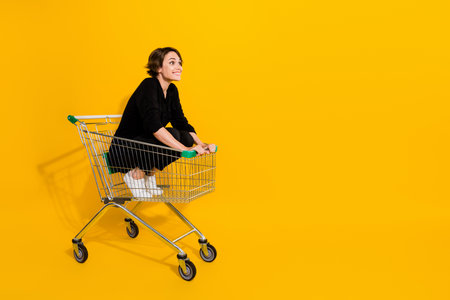 Photo of excited impressed lady wear black shirt riding shopping tray looking empty space isolated yellow color backgroundの写真素材
