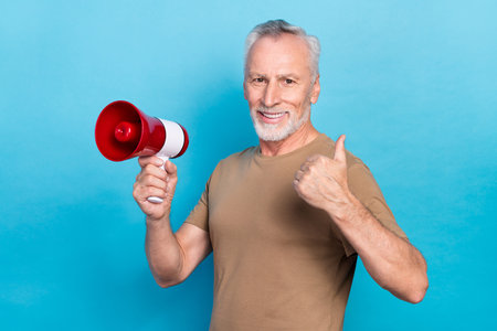 Photo of positive senior man gray hairstyle stubble wear beige t-shirt hold speaker showing thumb up isolated on blue color backgroundの写真素材