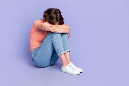 Full size photo of lovely young girl sit floor cuddle knees crying wear trendy striped garment isolated on violet color backgroundの写真素材