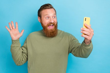 Portrait of positive cheerful guy with red hair wear khaki pullover say hi wave hand on video call isolated on turquoise color backgroundの写真素材