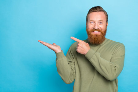 Portrait of satisfied guy with ginger hairdo wear khaki pullover palm indicating empty space isolated on turquoise color backgroundの写真素材