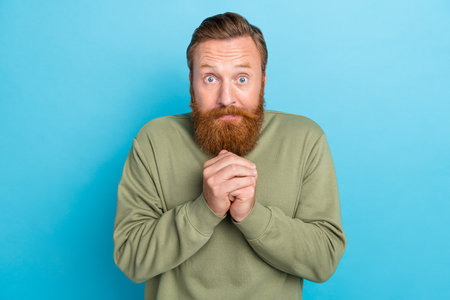 Portrait of pleading handsome man with red hairstyle dressed khaki pullover hold hands together isolated on blue color backgroundの写真素材