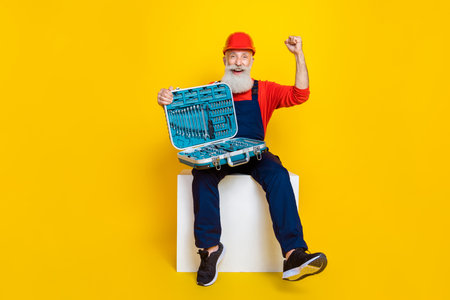 Photo of funny lucky age man wear overall uniform red hard hat rising fist open toolset isolated...