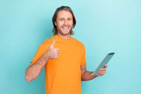Photo of confident cool man wear orange t-shirt reading modern device thumb up isolated turquoise...