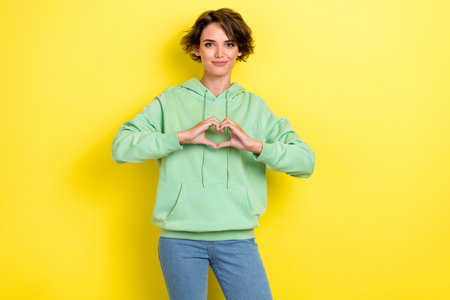 Photo portrait of lovely young woman showing heart fingers gesture dressed stylish green garment isolated on yellow color backgroundの写真素材