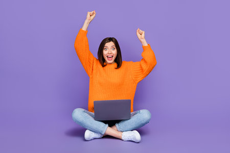 Full length photo of pretty young lady raise fists celebrate laptop dressed stylish orange knitted look isolated on purple color backgroundの写真素材