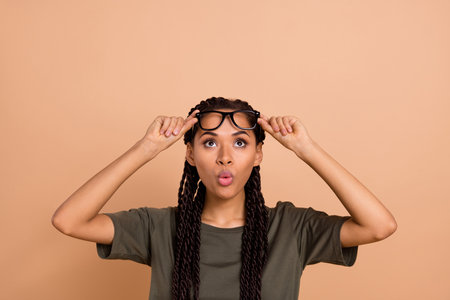 Portrait of impressed surprised girl hands touch eyeglasses look interested up empty space isolated on beige color backgroundの写真素材