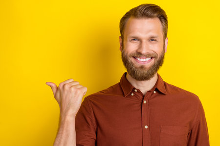 Photo of toothy beaming cheerful man with blond beard wear burgundy shirt directing empty space isolated on yellow color backgroundの写真素材