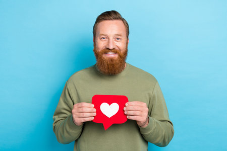 Portrait of glad good mood handsome guy with red hair wear khaki pullover hands hold red like isolated on turquoise color backgroundの写真素材