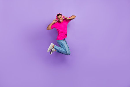 Full size photo of overjoyed cheerful man jumping hands touch headphones isolated on violet color...