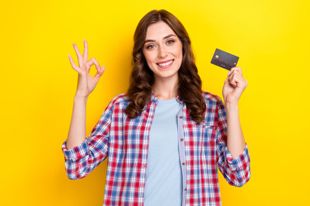 Photo of charming cute girl dressed checkered shirt rising debit card showing okey sign isolated yellow color backgroundの写真素材