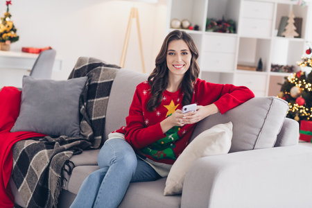 Photo of young satisfied pretty woman wear stylish winter outfit sitting her favorite coach living room relax with her smartphone stay homeの写真素材