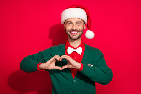 Photo of funky charming guy dressed xmas green cardigan showing arms heart celebrating noel isolated red color backgroundの写真素材