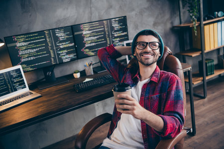 Photo of cheerful dreamy guy dressed eyewear smiling enjoying beverage arm hand behind head indoors workstation workshop homeの写真素材