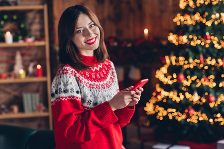 Photo portrait of young satisfied blogger girl wear ugly red sweater hold smartphone like eshopping before christmas holidays near fir tree indoorsの写真素材