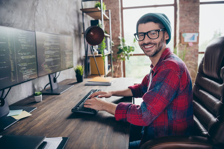 Photo of cheerful positive coder dressed hat glasses smiling writing system code indoors workplace...
